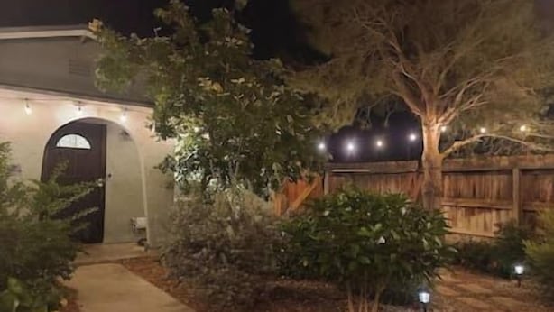 Una propiedad residencial con entrada pavimentada, cerco de madera y diversos arbustos y árboles.