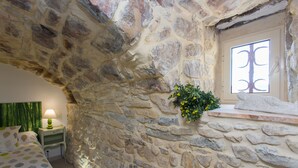 A stone-walled room with a bed, a nightstand, a lamp, and a window with a view.