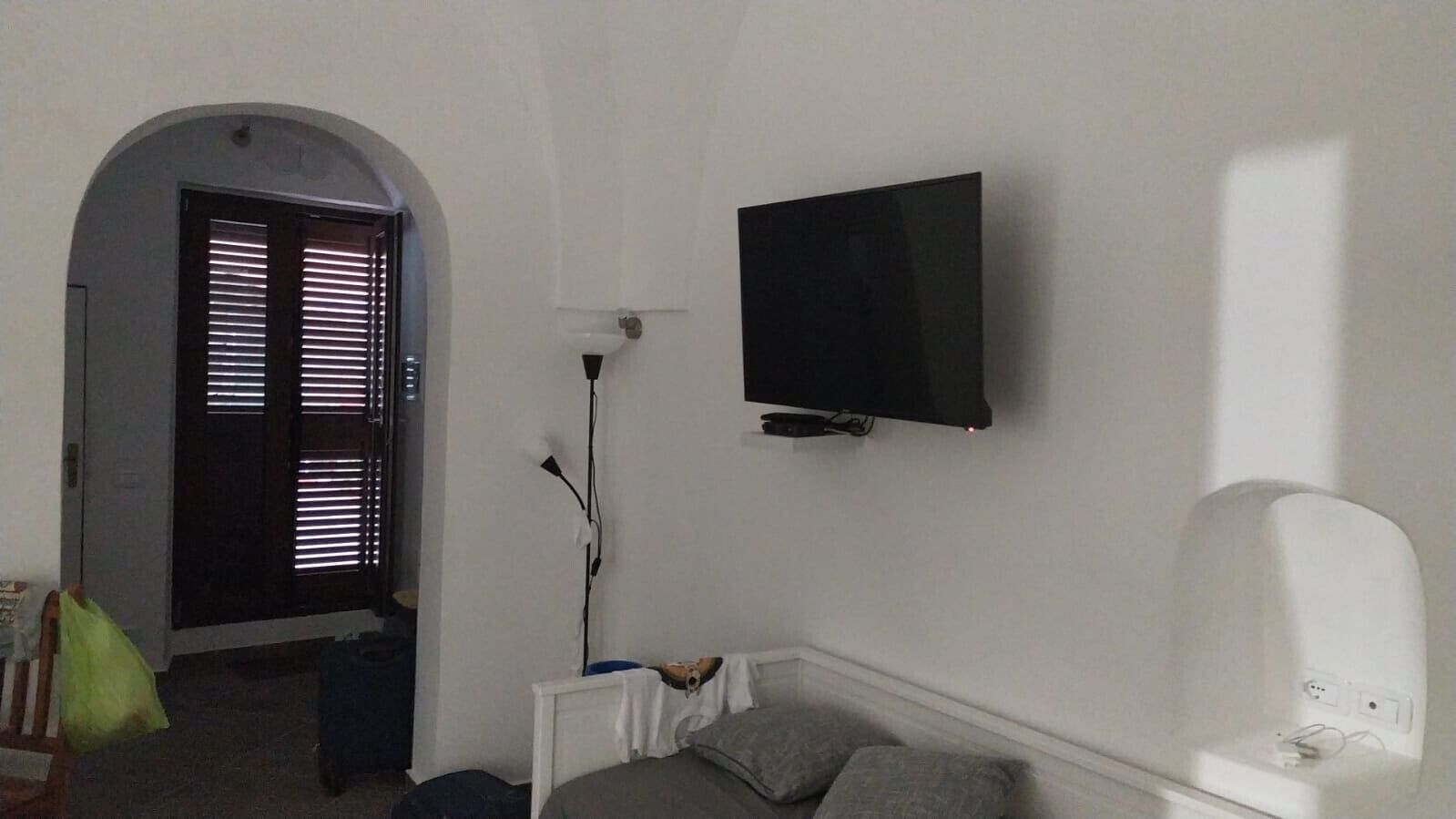 Een kamer met een aan de muur bevestigde televisie, een bed met kussens, een witte nachtkastje en een raam met zonwering.