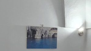 A room with a bed, a table with a patterned tablecloth, a wall-mounted picture of a coastal scene, and a ceiling light fixture. - Holiday Home "Villa Adriana" with Terrace & Wi-Fi (Pantelleria)