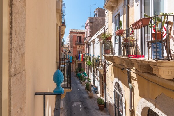 A narrow street with buildings on either side, featuring balconies with potted plants and a flag.