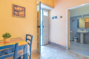 Une pièce comprenant une table, des chaises, une plante en pot, une porte donnant sur une salle de bain avec un lavabo et des murs carrelés.