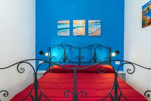 A bed with a red mattress in a room with blue walls, a metal headboard, and three paintings of the sea.