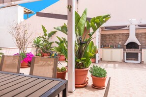 Een terras buiten met een tafel en stoelen, omgeven door potplanten en een bakoven.