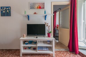 Un salon avec un téléviseur à écran plat posé sur un meuble blanc, une orchidée en pot et une étagère ornée d’objets décoratifs.