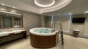 A modern bathroom with a wooden bathtub, two sinks, a large mirror, and a flat-screen TV mounted on the wall. - Golden Tulip Jalandhar (Jalandhar)