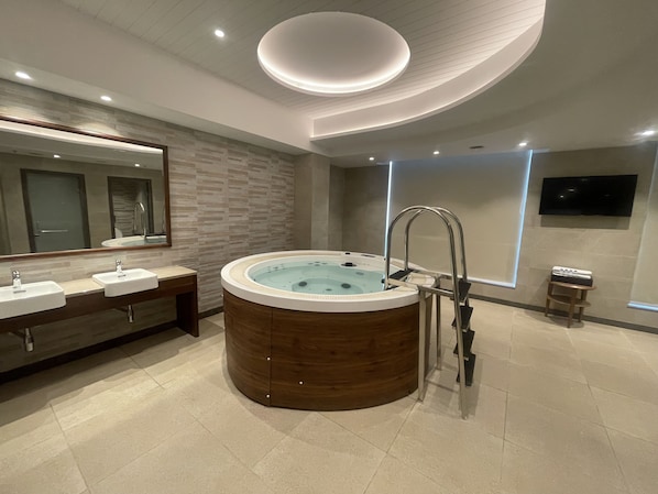 A modern bathroom with a wooden bathtub, two sinks, a large mirror, and a flat-screen TV mounted on the wall.
