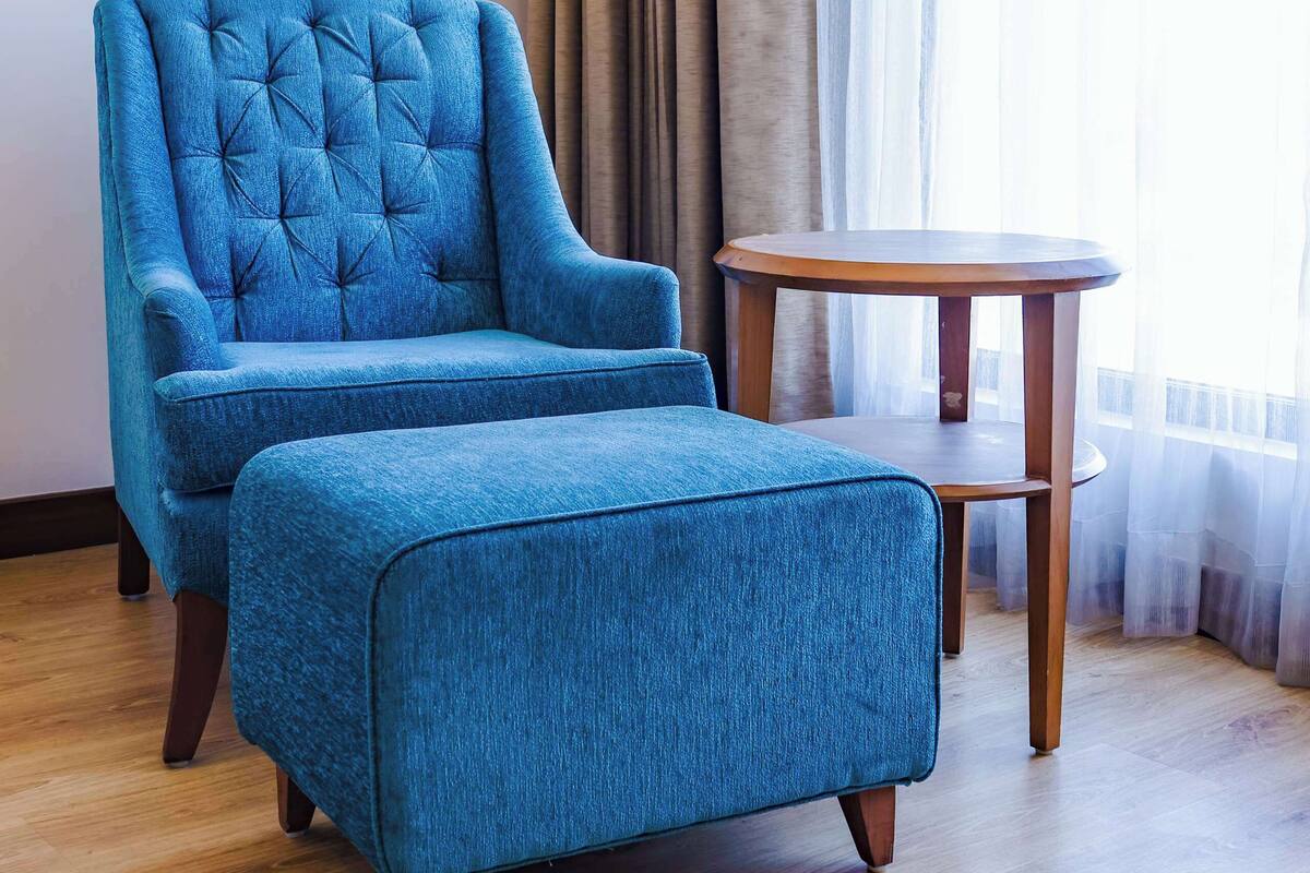 Ein Zimmer mit einem gesteppten blauen Sessel und Hocker, einem runden Holztisch und einem großen Fenster mit transparenten Vorhängen.