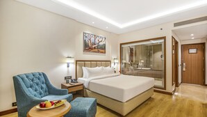 A hotel room with a large bed, a blue armchair, a small table with fruit, a painting on the wall, and a glass-enclosed shower area. - Golden Tulip Jalandhar (Jalandhar)