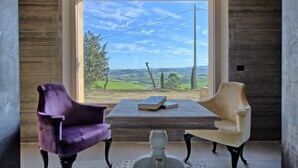 Een kamer met een groot raam dat uitzicht biedt op groene velden en heuvels. Er staan twee stoelen en een tafel met een boek erop.