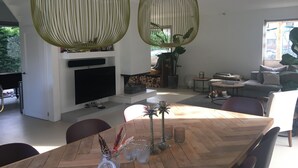 A modern living room with a large TV, wooden dining table, and hanging green lamps. - Beautiful villa with sauna, 10 min from the beach (Bentveld)