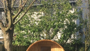 A wooden barrel structure with a door and two benches, surrounded by a tree and a fence. - Beautiful villa with sauna, 10 min from the beach (Bentveld)