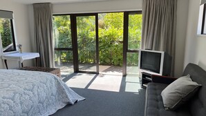 Una habitación de hotel moderna con una cama grande, un sofá gris, un televisor y un ventanal corredero que da a un balcón con vegetación.