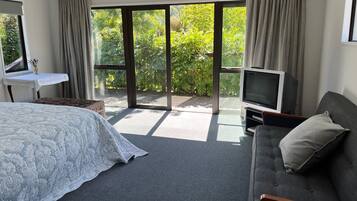 A modern hotel room with a large bed, a grey sofa, a television, and a sliding glass door leading to a balcony with greenery.