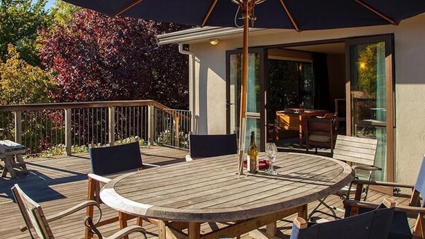 Una terraza de madera con una mesa redonda y sillas, una sombrilla y una zona de bar con cerramiento de vidrio.