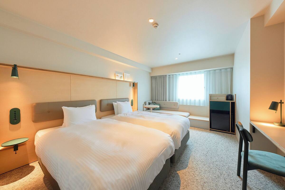 A hotel room with two beds, a desk, a chair, a TV, and a window with curtains.