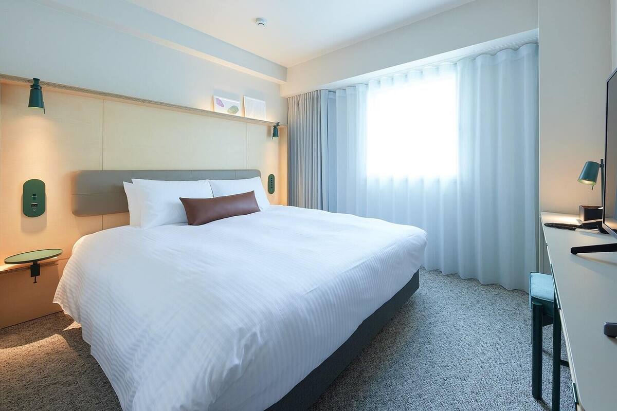 A hotel room with a large bed, a desk with a flat-screen TV, a small table, and a wall-mounted control panel.