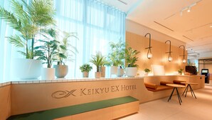 A reception area with a Keikyuu Ex Hotel sign, a green bench, a seating area with a sofa and two tables, and potted plants. - Keikyu EX Hotel Sapporo (Sapporo)
