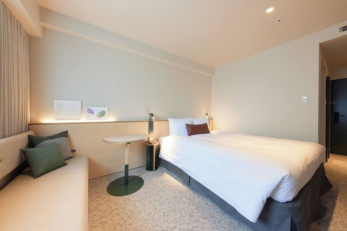 A hotel room with a bed, a small bedside table, a lamp, a chair, and a wall-mounted artwork.