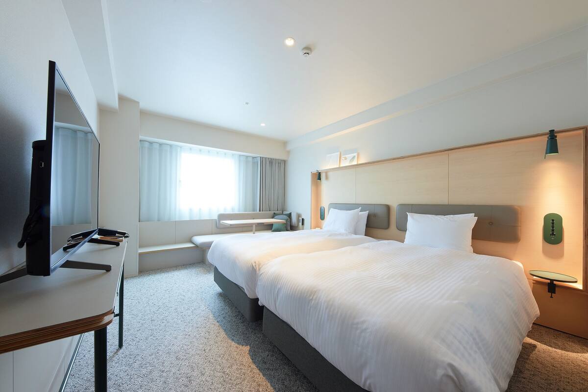 A hotel room with a large bed, a desk with a flat-screen TV, a seating area with a bench, and a window with curtains.
