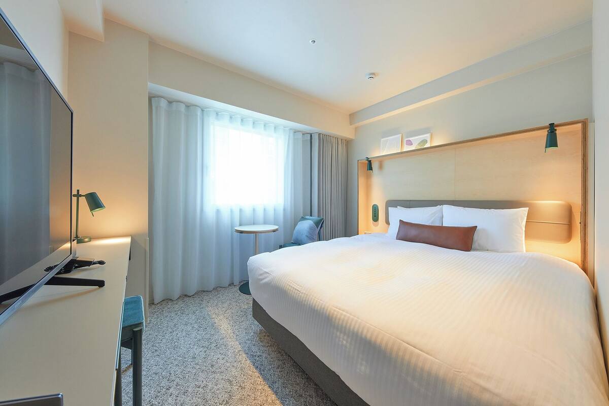 A hotel room with a large bed, a desk with a flat-screen TV, a small table, and a window with curtains.