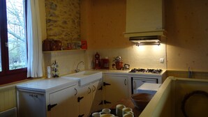 A compact kitchen with a stone wall, wooden cabinets, a sink, a stove, and a window with curtains. - Gîtes Pyrénées Les Trois Montagnes BALOU (Ganties)