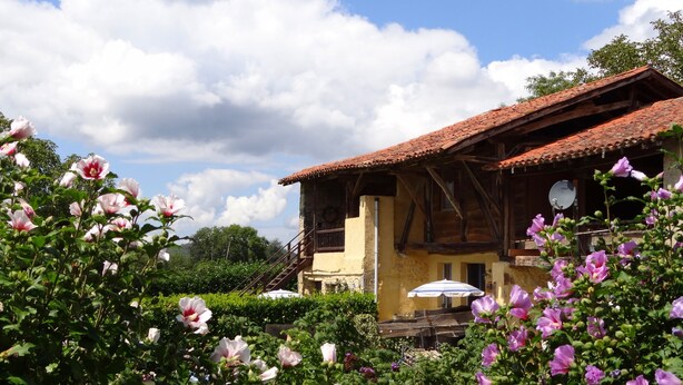 A rustic house with a tiled roof, surrounded by lush greenery and blooming flowers. - Gîtes Pyrénées Les Trois Montagnes BALOU (Ganties)