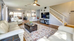 A spacious living room with a white leather sofa, a wooden coffee table, and a large rug. A ceiling fan and a TV are mounted on the wall.