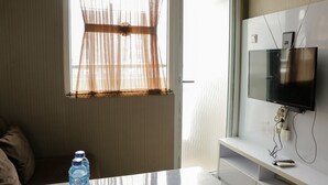 A hotel room with a large window, a flat-screen TV mounted on the wall, a white table, and a water bottle on the table. - Homey And Cozy Stay 2Br At Green Pramuka City Apartment (Jakarta)