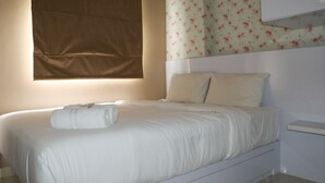 A neatly made bed with white linens and a folded towel, a window with a brown blind, and a wall with floral wallpaper. - Homey And Cozy Stay 2Br At Green Pramuka City Apartment (Jakarta)
