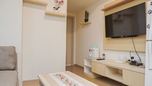A living room with a sofa, a coffee table with a floral table runner, a TV mounted on the wall, a small shelf with plants, and a desk with a water dispenser. - Comfort 1Br With Working Room At Meikarta Apartment (Cikarang)