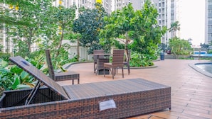 A wicker lounge chair with a tag, a wicker table, and chairs in a landscaped outdoor area with a pool. - Fancy And Nice Studio Apartment At Springlake Summarecon Bekasi (Bekasi)