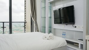 A hotel room with a bed, a flat-screen TV mounted on the wall, a window with a view of the city, and a water bottle on the nightstand. - Homey And Warm Studio At Sky House Bsd Apartment (Pagedangan)