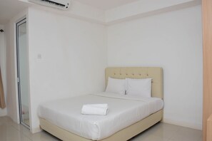 A neatly made bed with a beige headboard and white bedding in a room with white walls and a sliding glass door. - Cozy And Strategic Studio At Bassura City Apartment (Jakarta)