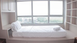 A bed in a corner with a large window, built-in shelving, and a view of buildings and trees. - Warm And Cozy Studio At Springlake Summarecon Apartment (Bekasi)