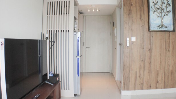 A compact living space with a television, a refrigerator, and a wooden wall with a tree painting. - Warm And Cozy Studio At Springlake Summarecon Apartment (Bekasi)