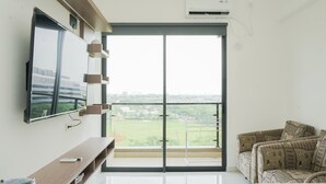 A modern living room with a large TV, a sofa, and a balcony with a city view. - Homey And Cozy Living 2Br At Sky House Bsd Apartment (Pagedangan)