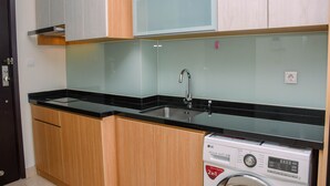 A compact kitchen with wooden cabinets, a black countertop, a stainless steel sink, and a front-loading washing machine. - Great Deal And Comfy Studio At Menteng Park Apartment (Jakarta)