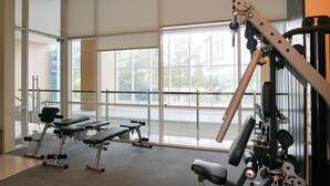 Een sportschool met gewichthefapparatuur en banken, grote ramen en uitzicht op bomen en gebouwen.