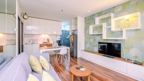 A modern living room with a sofa, coffee table, and a TV. There is a kitchen area with cabinets and appliances in the background.