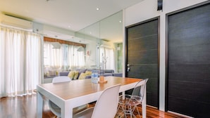 A modern dining area with a wooden table and white chairs, a kitchen in the background, and a large dark door.