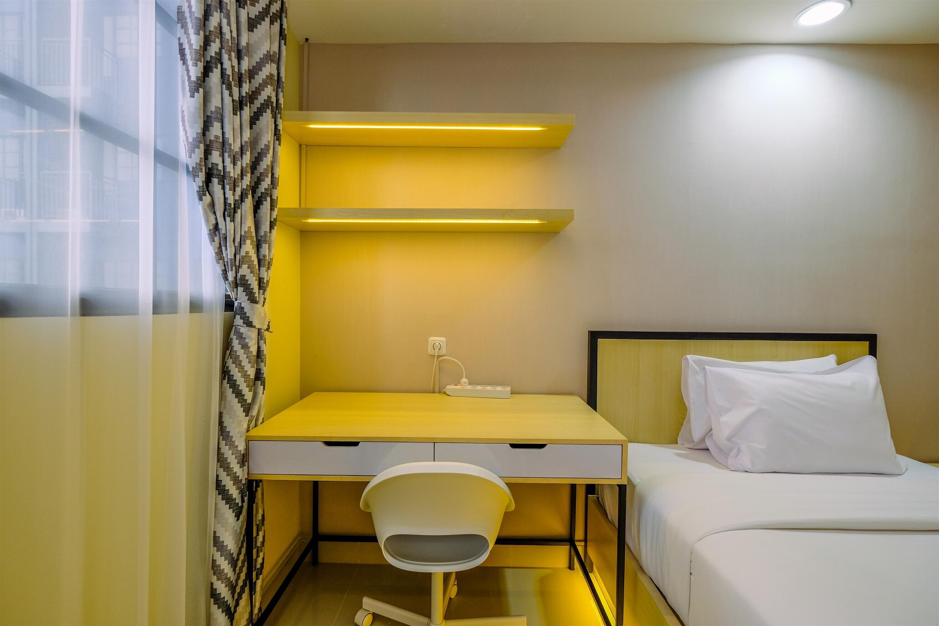 A compact hotel room with a bed, a desk with a chair, and a small window with patterned curtains.