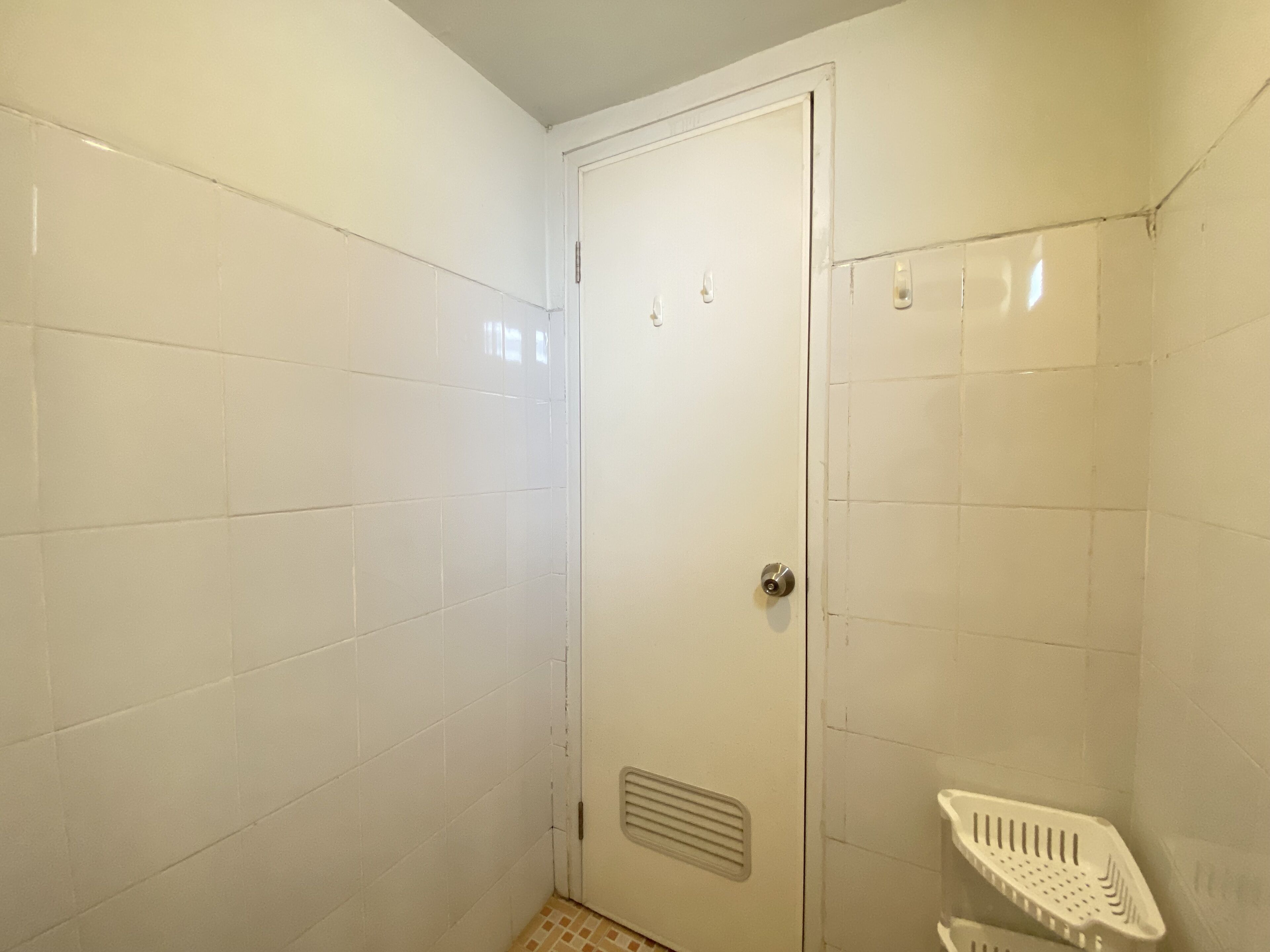 Une petite salle de bain aux murs recouverts de carreaux blancs, avec une zone douche, une porte et une étagère en plastique blanche.