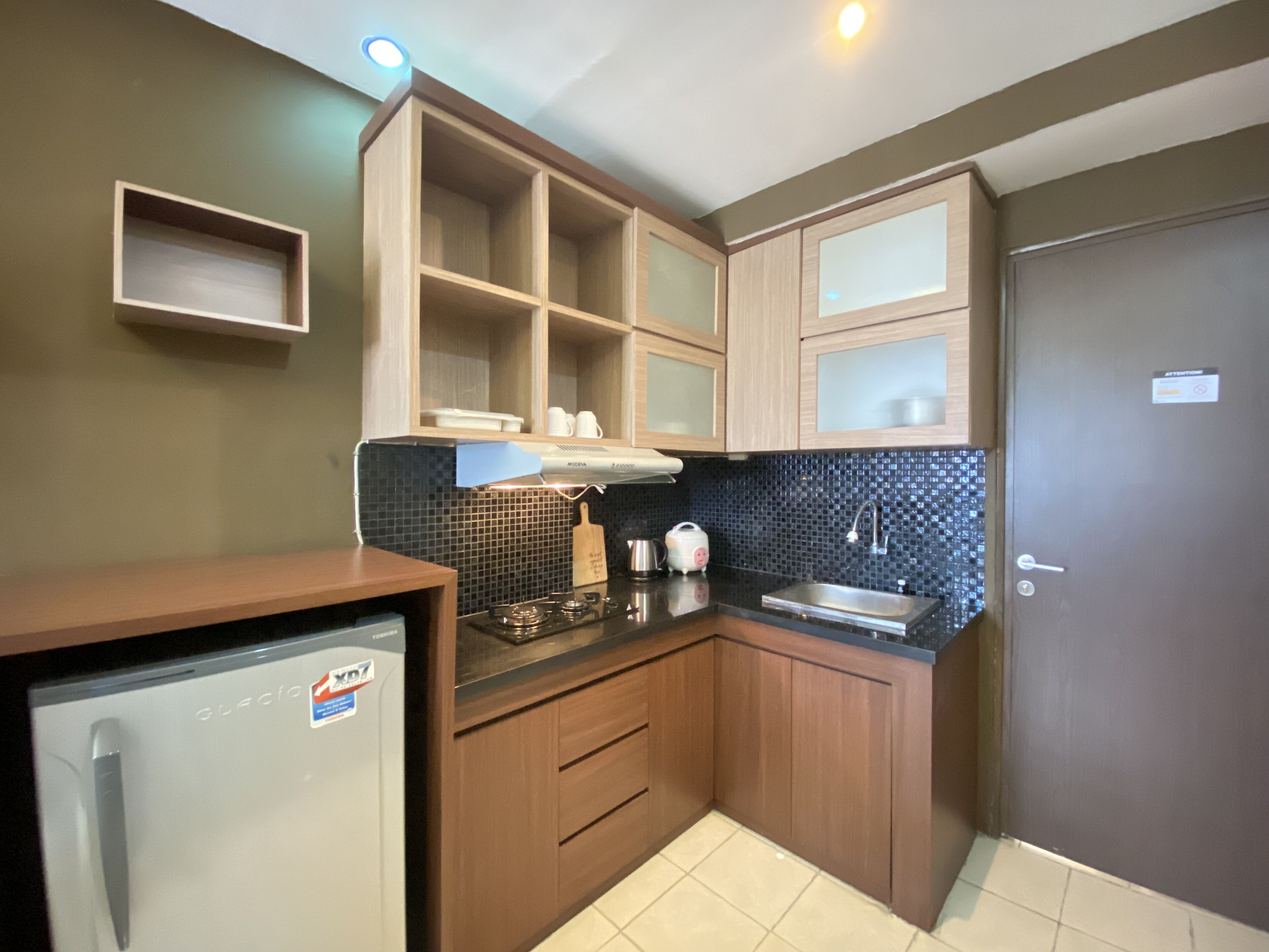 A compact kitchen with wooden cabinets, a black tiled backsplash, a stainless steel sink, and a refrigerator.