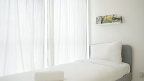 Un llit ben arrenjat amb llençols blancs i un panell de capçal gris a una habitació lluminosa amb cortines blanques.