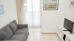 Un saló modern amb un sofà gris, un televisor de pantalla plana sobre un suport blanc i una finestra gran amb cortines transparents.