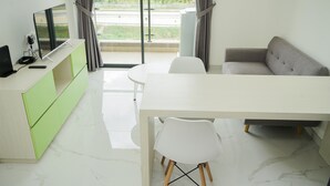 A modern living room with a flat-screen TV, a sofa, a dining table, and a view of greenery through large windows. - Fully Furnished With Modern Design 2Br At Sky House Bsd Apartment (Pagedangan)