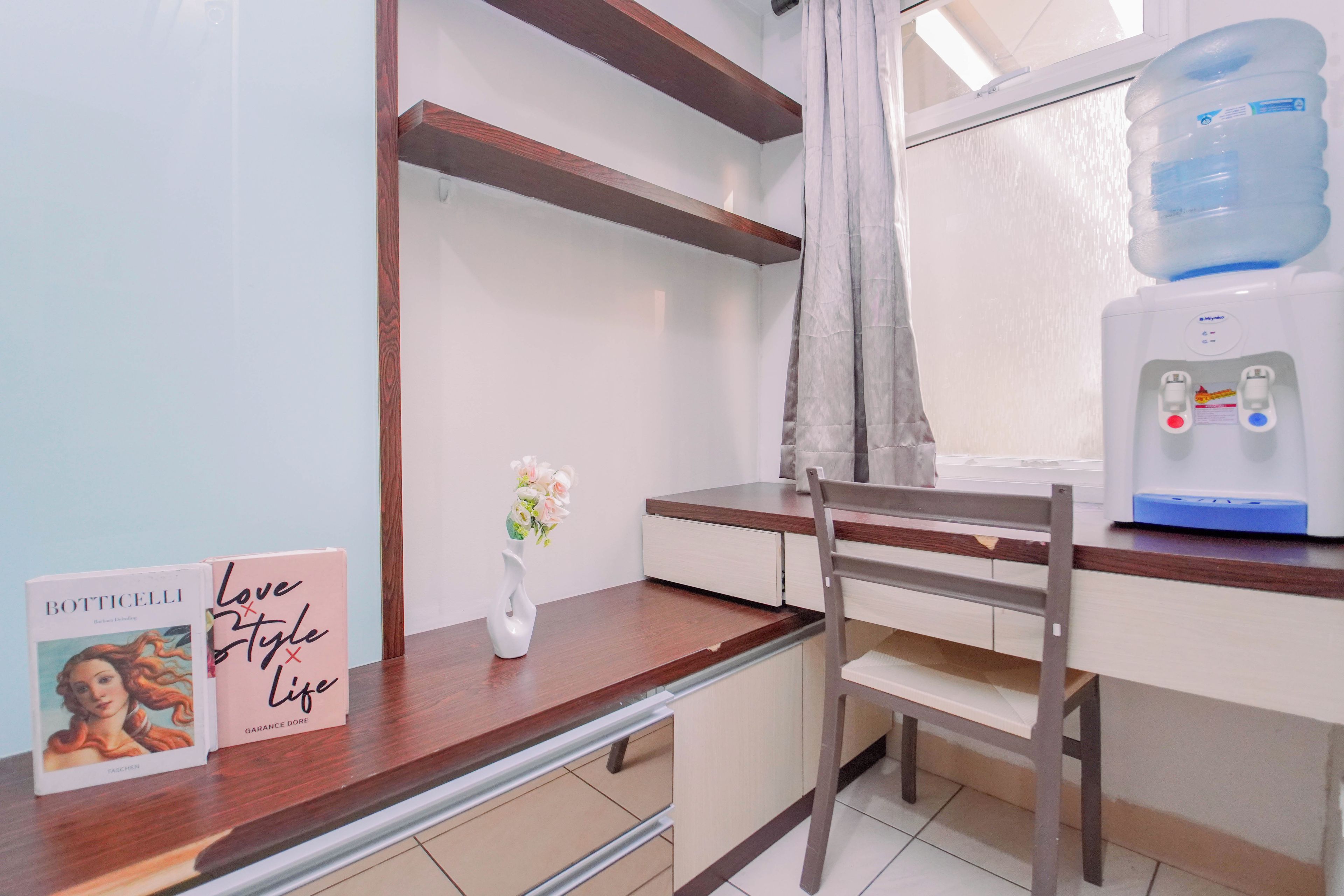 A hotel room with a wooden desk, a water dispenser, a chair, and a vase with flowers.
