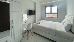 A bedroom with a bed, a desk, a chair, a TV, and a window with a city view. - Bright And Relaxing 2Br At Mekarwangi Square Bandung (Bandung)