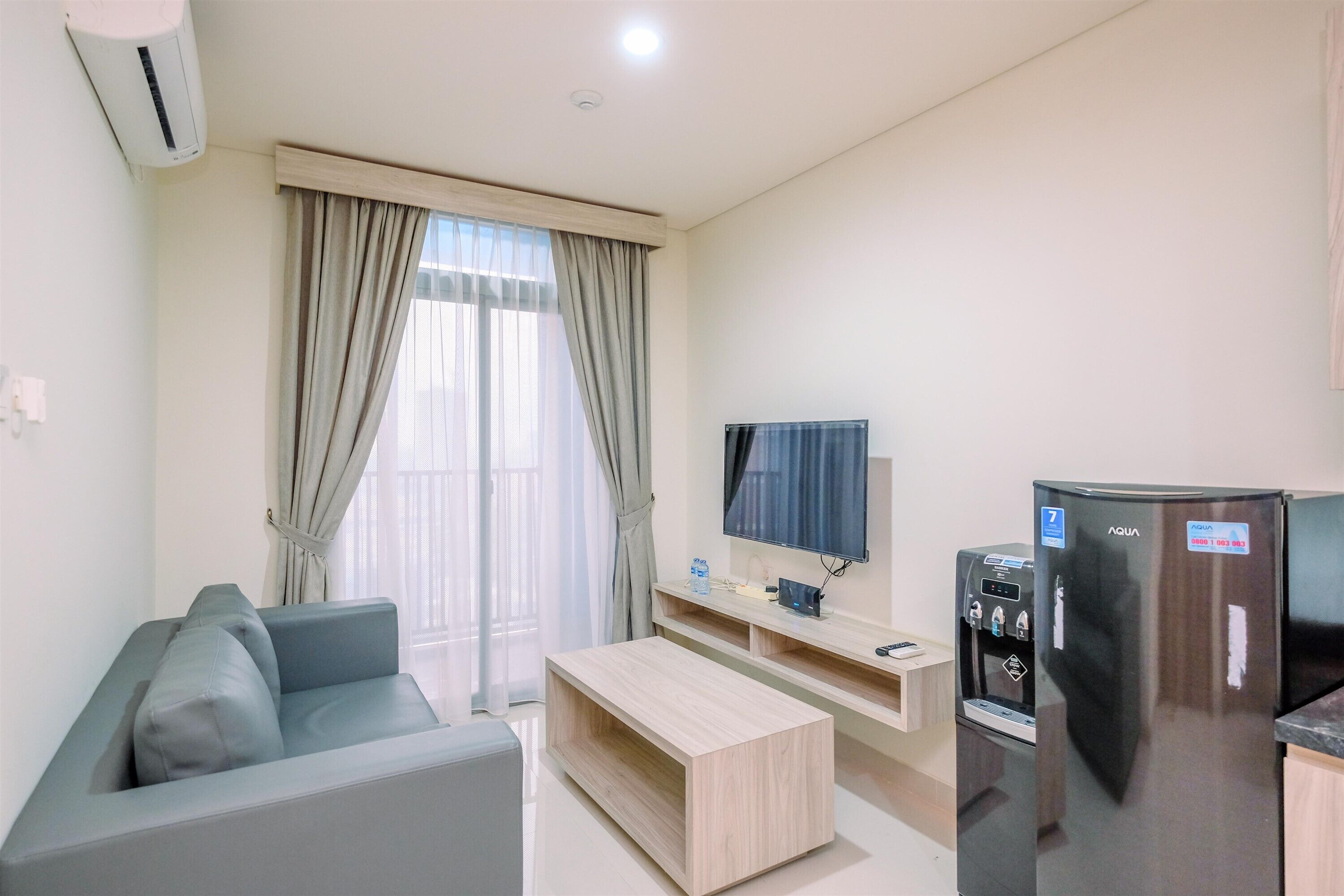 Una sala de estar moderna con un sofá gris, una mesa de centro de madera, un televisor de pantalla plana, un refrigerador y un dispensador de agua.
